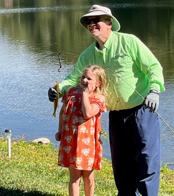 Tom in green shirt and hat holding fishing pole and fish with young girl in pink.