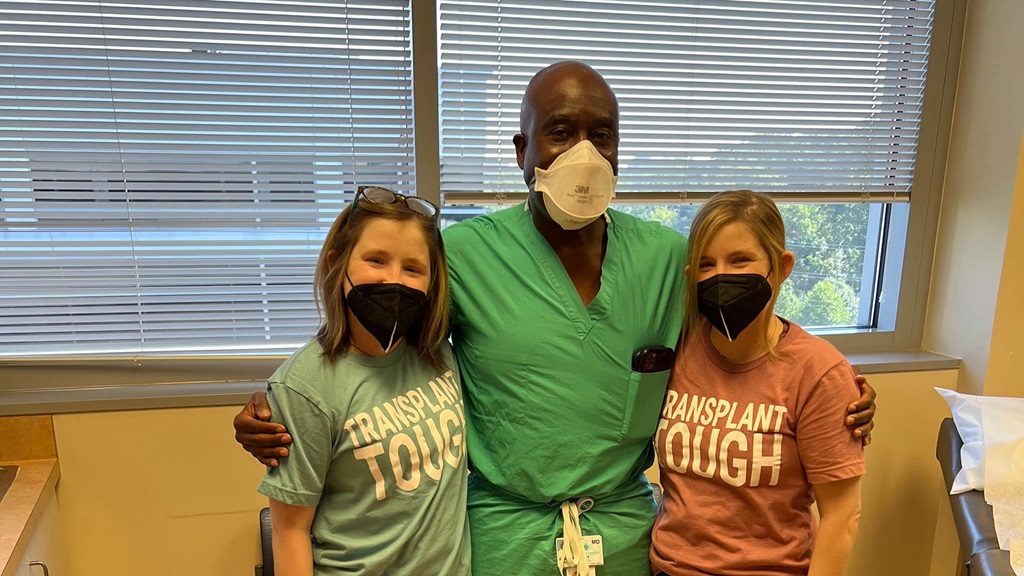 Twins in blue and pink shirts pose for a photo with their doctor. All three are wearing masks.