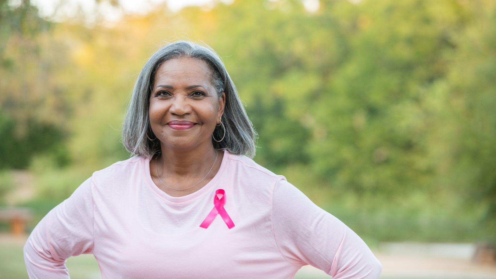 Woman wearing pink shirt with pink breast cancer ribbon, smiling in front of green foliage.