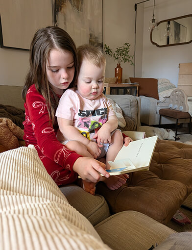 An infant girl and a young girl sitting together and reading a book.