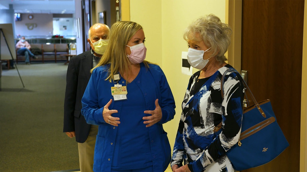 Two women wearing masks, one in blue scrubs and one in a blue and white shirt talking and touring a medical facility.
