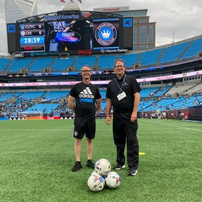 Two men from the Charlotte Football club, smiling inside the Charlotte soccer stadium with soccer balls at their feet.