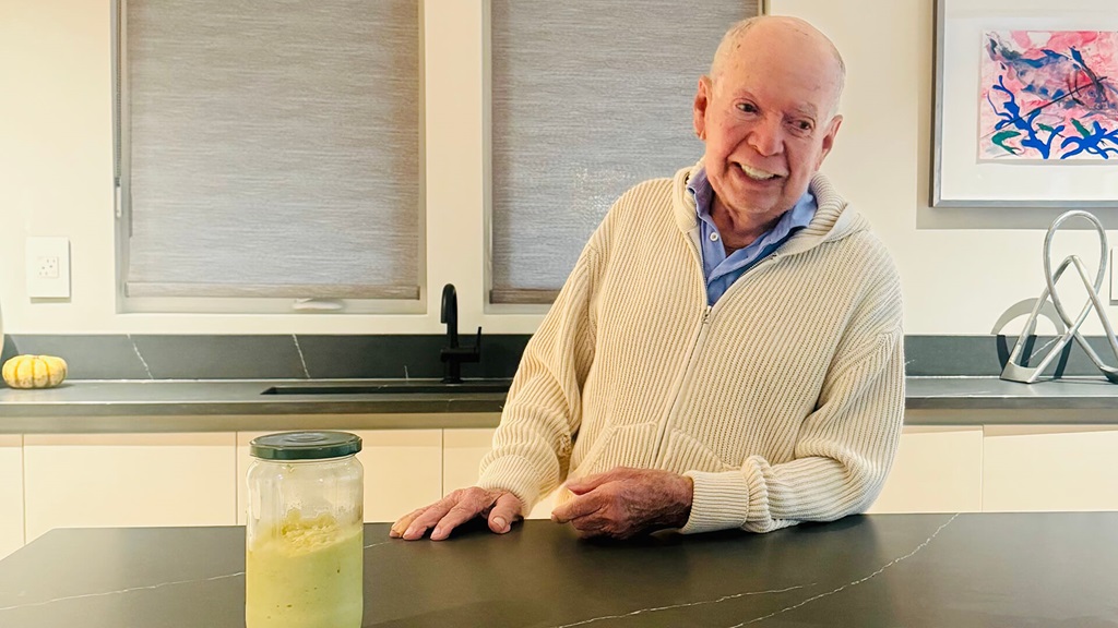An old man in a white sweater smiling and sitting at a table.