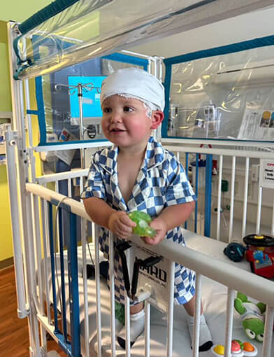 A young boy standing in a crib while wearing a bandage on his head and a blue checkered outfit.