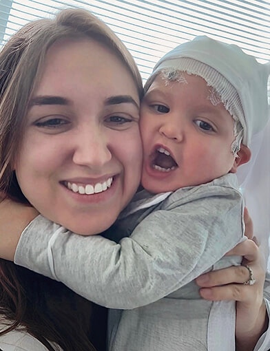 A young boy in a gray hat and shirt being held by his mother.