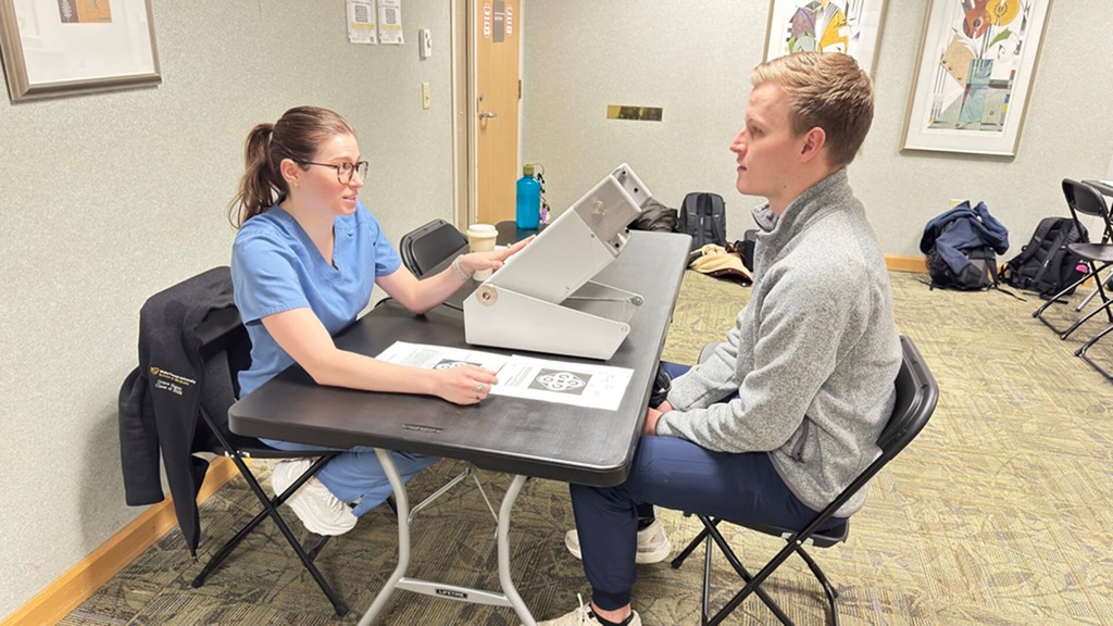 A young man sitting in a chair taking a vision test administered by a young woman in blue scrubs.