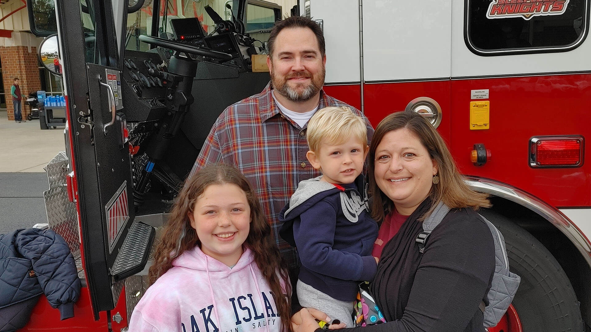 A family of four smiling at the camera, standing in front of a fire truck.