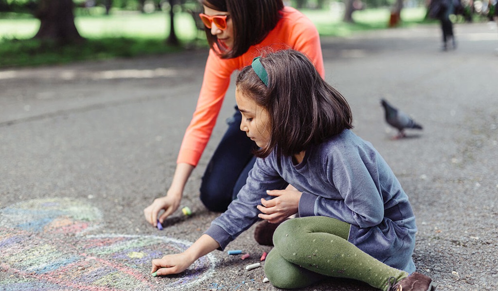 A young adult and a child in long sleeve shirts and pants drawing on asphalt with chalk.