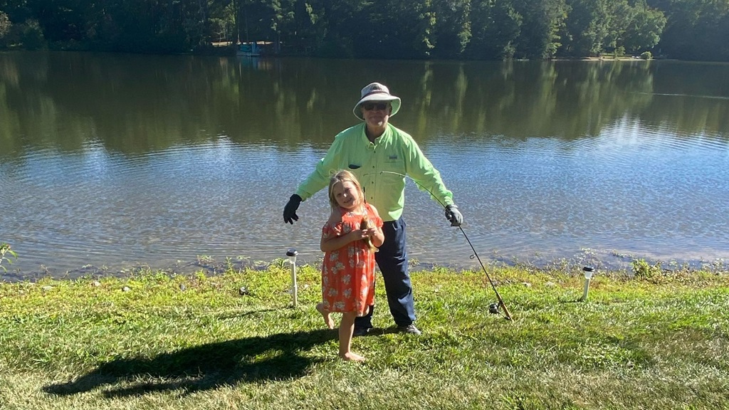 Tom wearing green jacket holding fishing poles and fish in front of water on grass with young girl in pink.