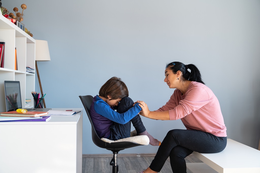 Child sitting on chair with head palming legs and arms folded, parent consoling 