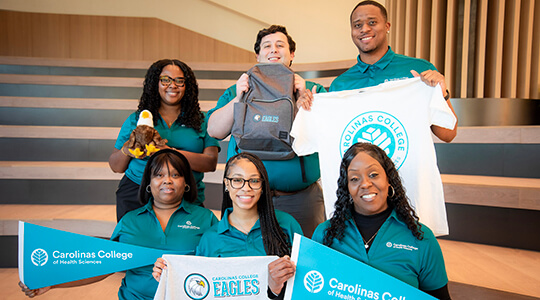 A group of women and men wearing teal shirts standing together in a group and holding pennants, shirts, bags and stuffed animals.