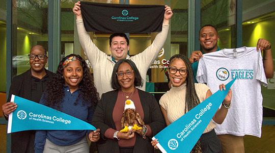 A group of men and women stand for a group photo holding teal pennants for Carolinas College of Health Science.
