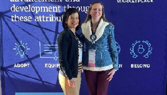 Two women in business casual attire standing and smiling in front of a backdrop with information about the Sigma Marketplace on it.