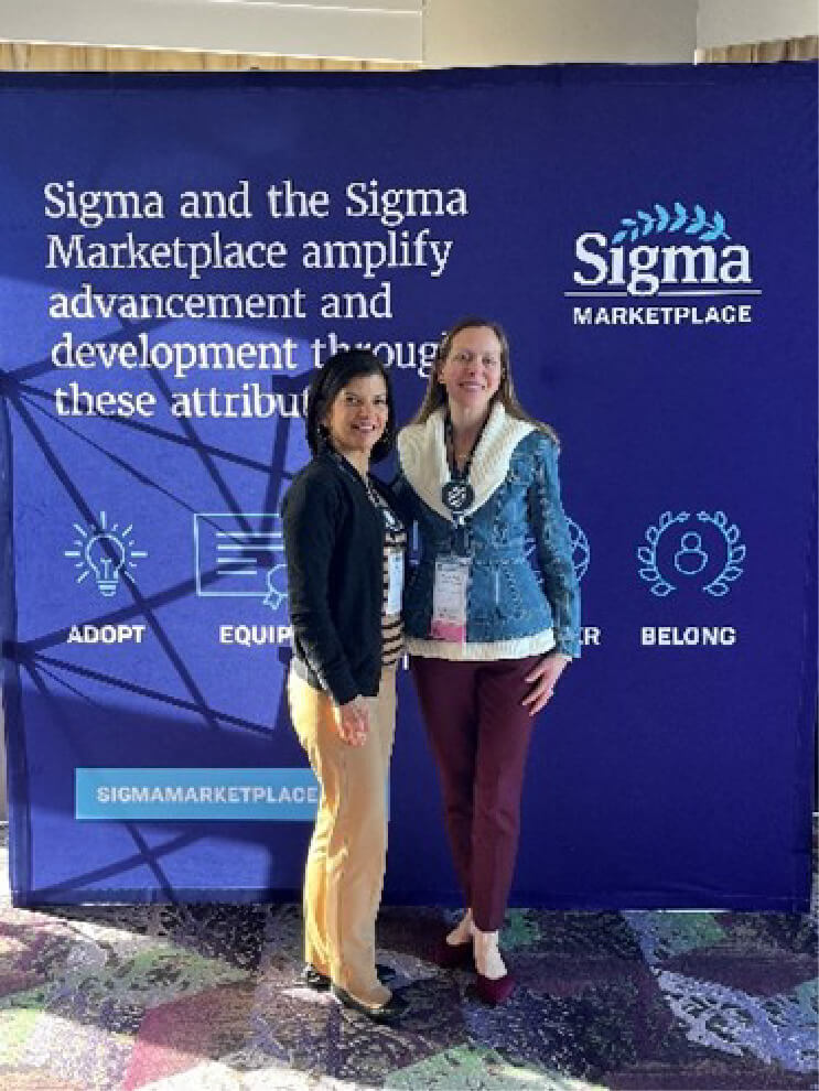 Two women in business casual attire standing and smiling in front of a backdrop with information about the Sigma Marketplace on it.