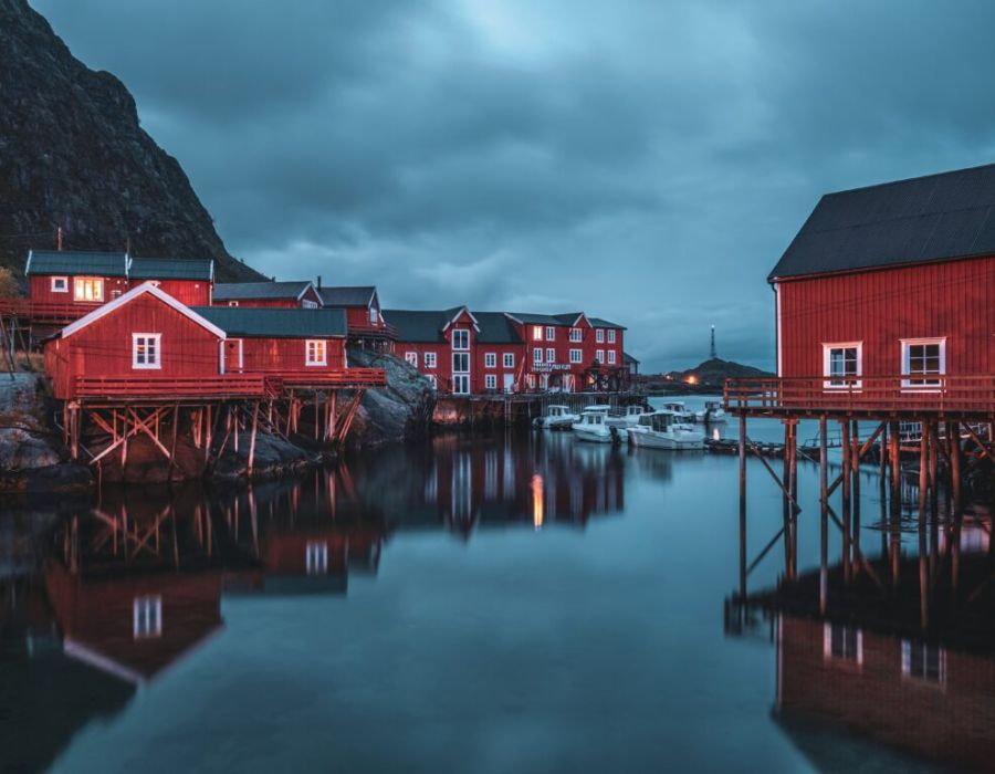 Red houses on pylons above water at nighttime with cloudy overcast sky.