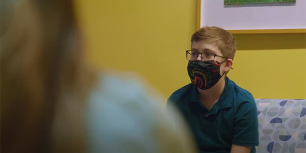 A young boy with brown hair wearing glasses and a mask sits in a room with yellow walls to receive music therapy.